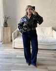 Person wearing a dark brown leather jacket and blue jeans taking a mirror selfie in a room with a white couch and wooden side table.