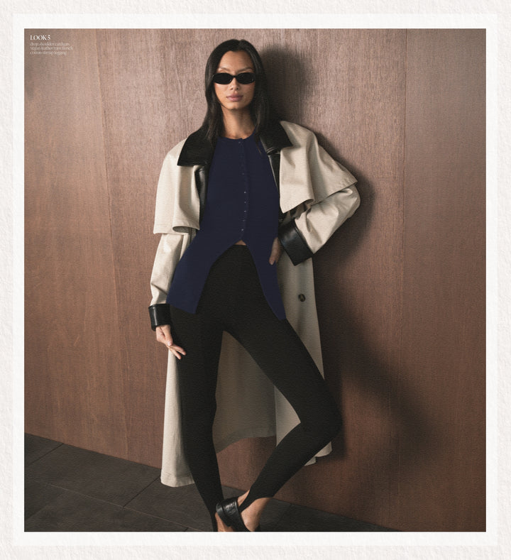 Woman in a trench coat and sunglasses posing against a wooden wall.