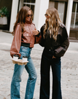 Two women standing outdoors on a cobblestone street, each wearing a cropped leather jacket. The woman on the left wears a brown leather jacket with blue jeans and holds a coffee cup carrier.